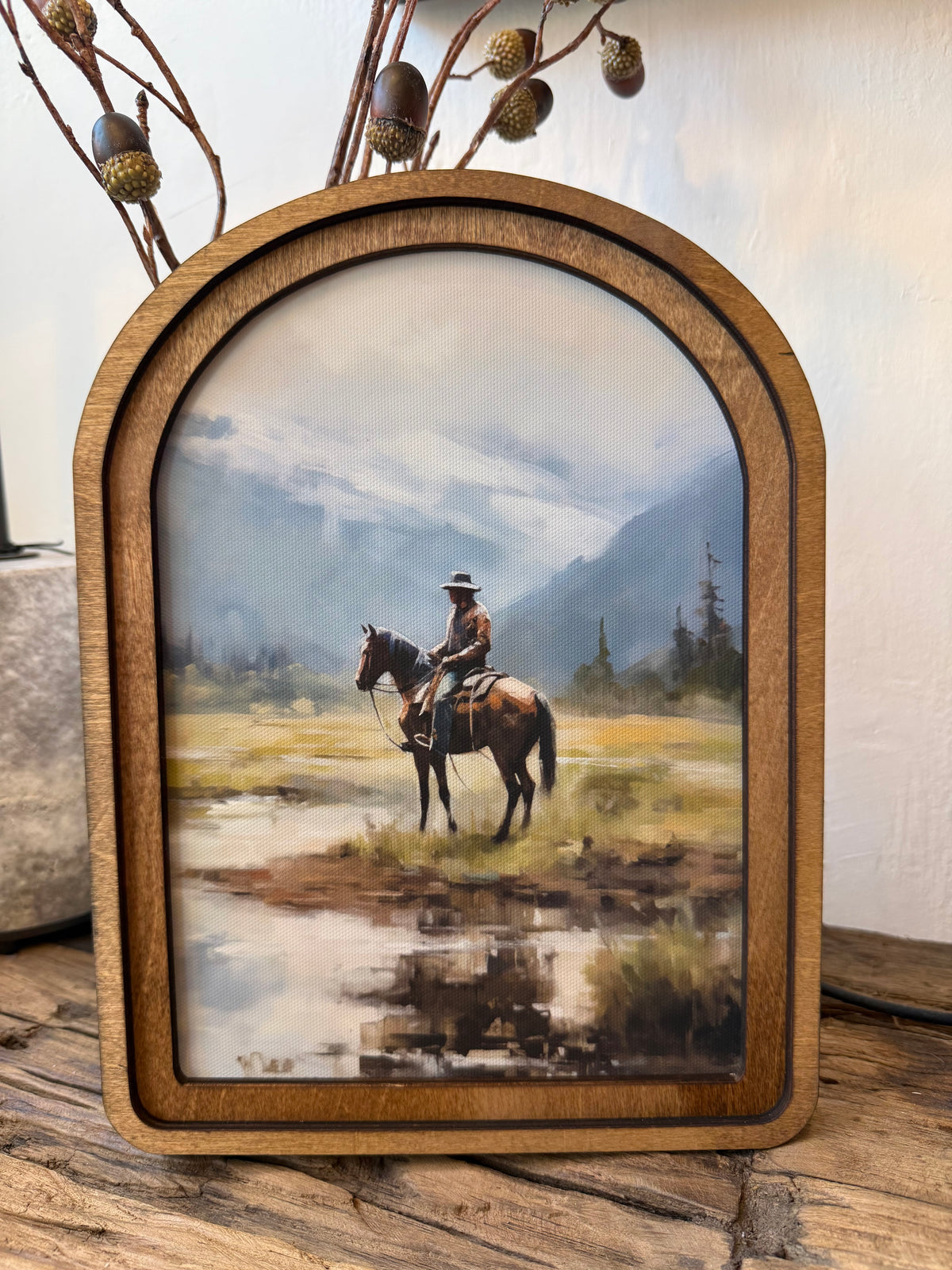 An arched wooden picture frame holding a western landscape painting of a cowboy riding a horse through a marshy field with mountains in the background, displayed on a rustic wooden surface.