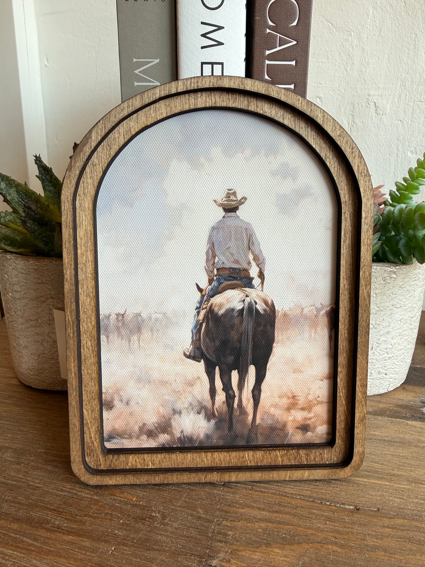 A wooden arched picture frame with a rustic finish, displaying a western-themed print of a cowboy riding a horse through dust. The frame is styled on a wooden tabletop with potted plants and books in the background.