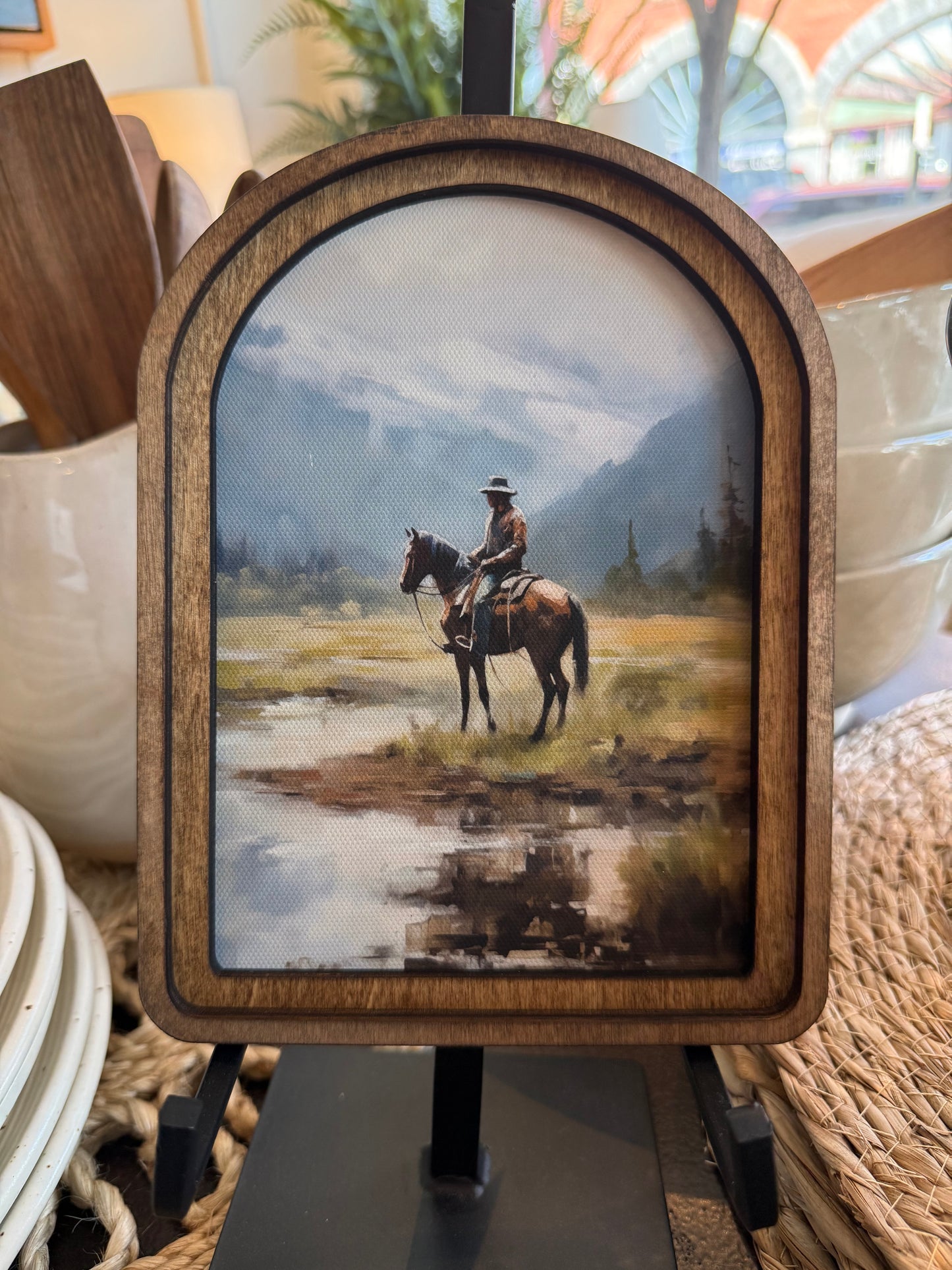 A framed landscape art print featuring a cowboy riding a horse through a marshy field with mountains in the background, displayed on a black stand.