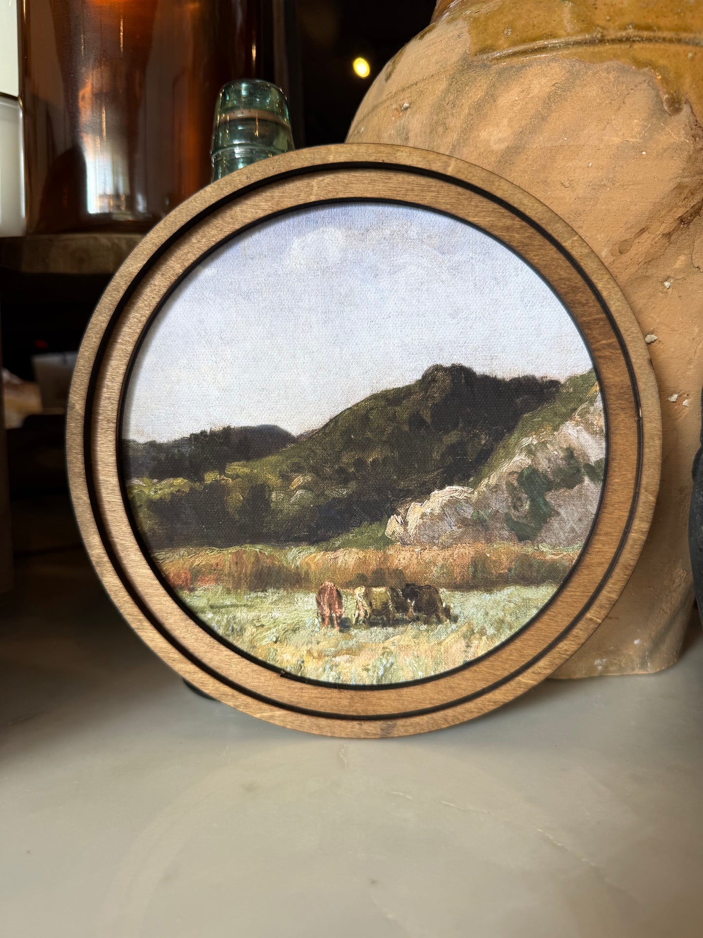 A round framed landscape painting depicting a pastoral field with cows, hills, and a pale sky, set in a rustic wooden barrel or container on a tabletop.