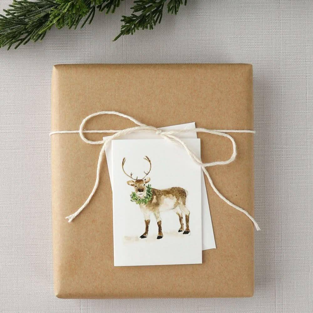 A square gift box wrapped in brown kraft paper and tied with a white string bow, featuring a small white card with a watercolor illustration of a deer wearing a green wreath. Pine branches are visible at the top edge against a light textured fabric background.