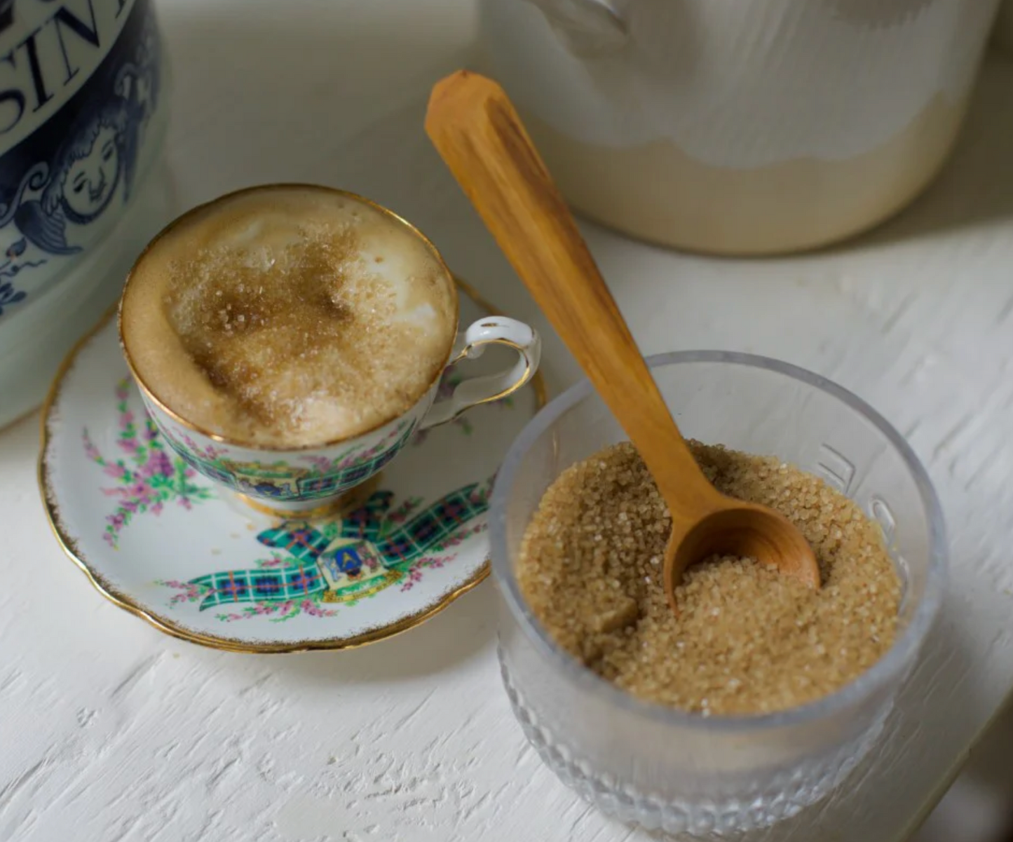 Cappuccino with a side of brown sugar in a glass bowl on a white surface.