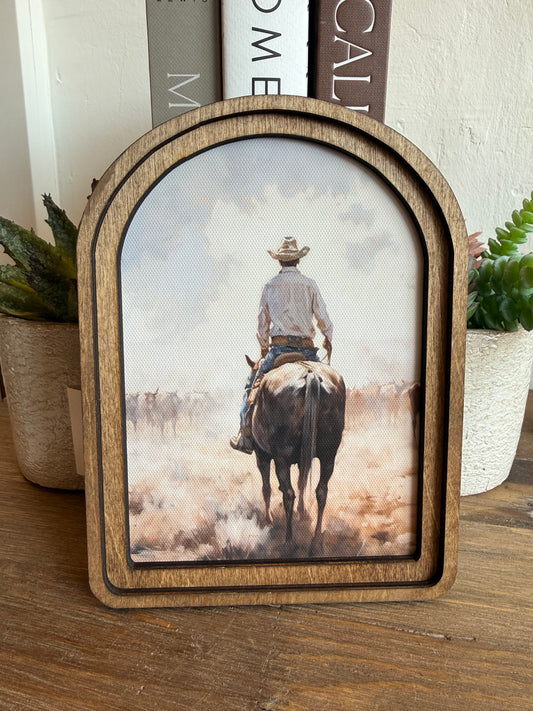 A wooden arched picture frame with a rustic finish, displaying a western-themed print of a cowboy riding a horse through dust. The frame is styled on a wooden tabletop with potted plants and books in the background.