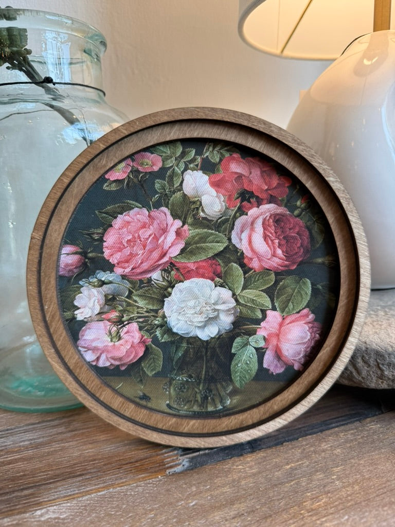 Round wooden framed floral print featuring pink and white roses with green leaves, styled on a wooden surface next to a glass jar and a lamp.