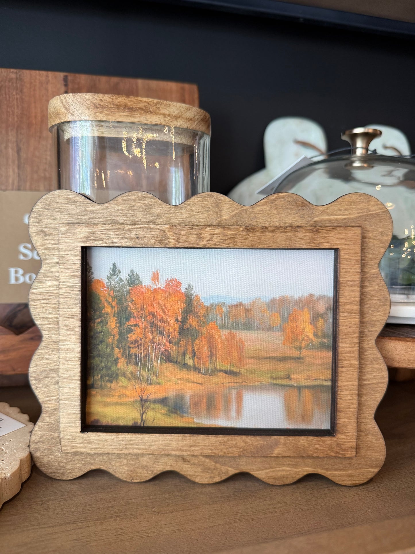 A scalloped wooden picture frame with a natural oak finish, holding a landscape print of an autumn lake scene, displayed on a wooden shelf next to a glass jar.