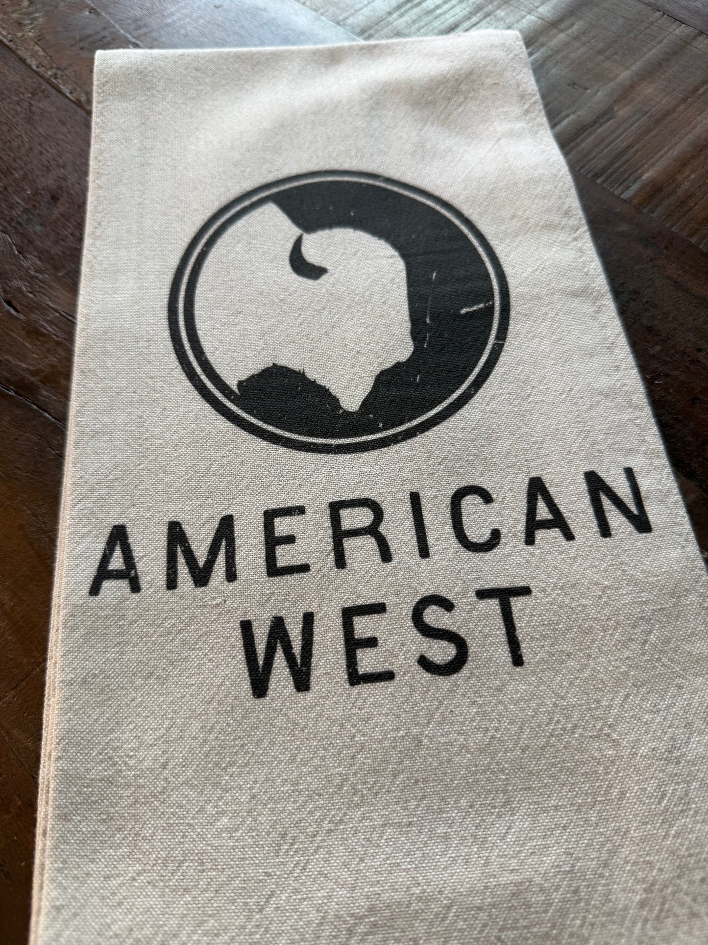 Close-up of the American West tea towel showcasing the natural stonewashed fabric, hemmed edges, and printed logo.
