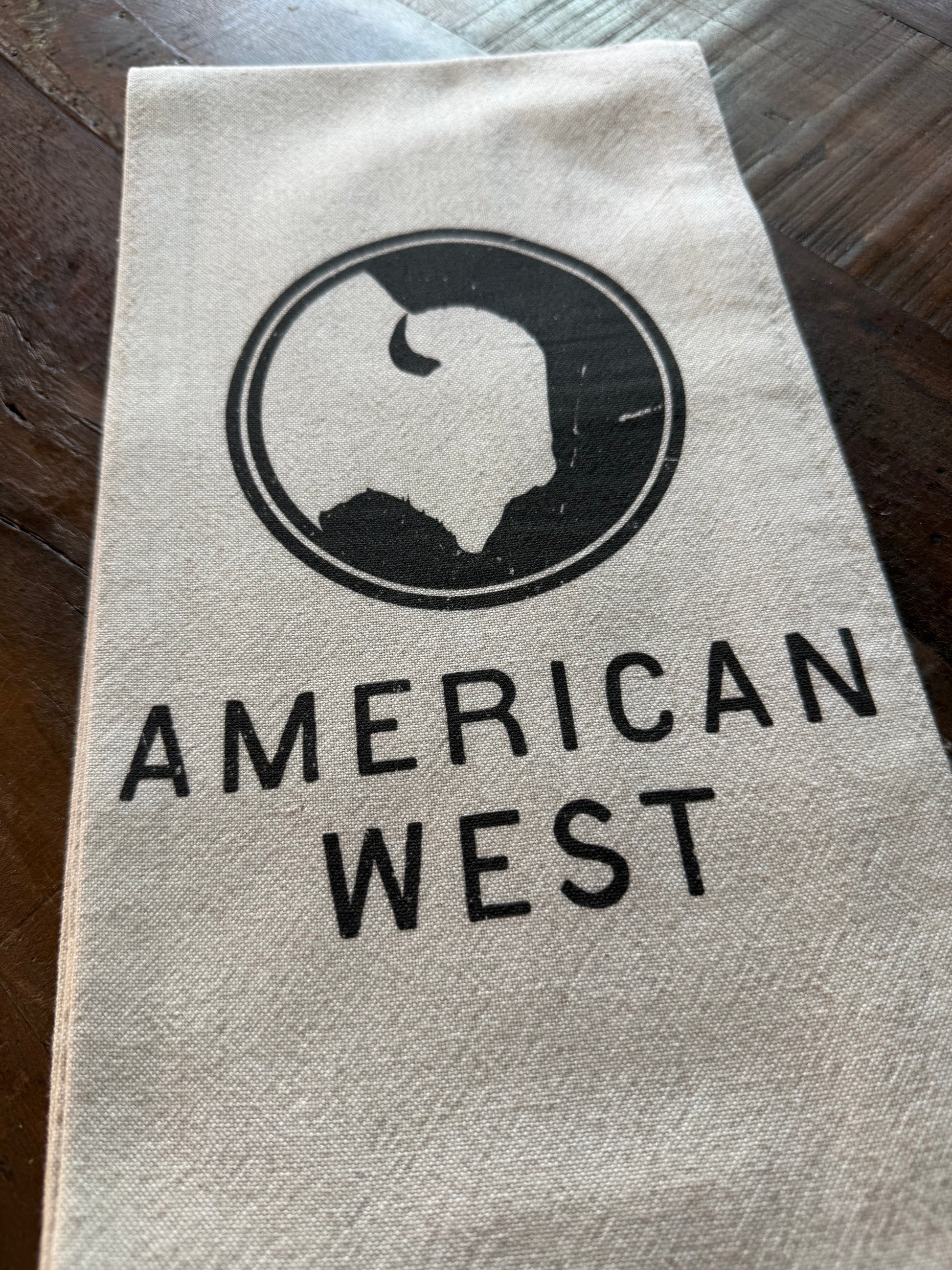 Close-up of the American West tea towel showcasing the natural stonewashed fabric, hemmed edges, and printed logo.