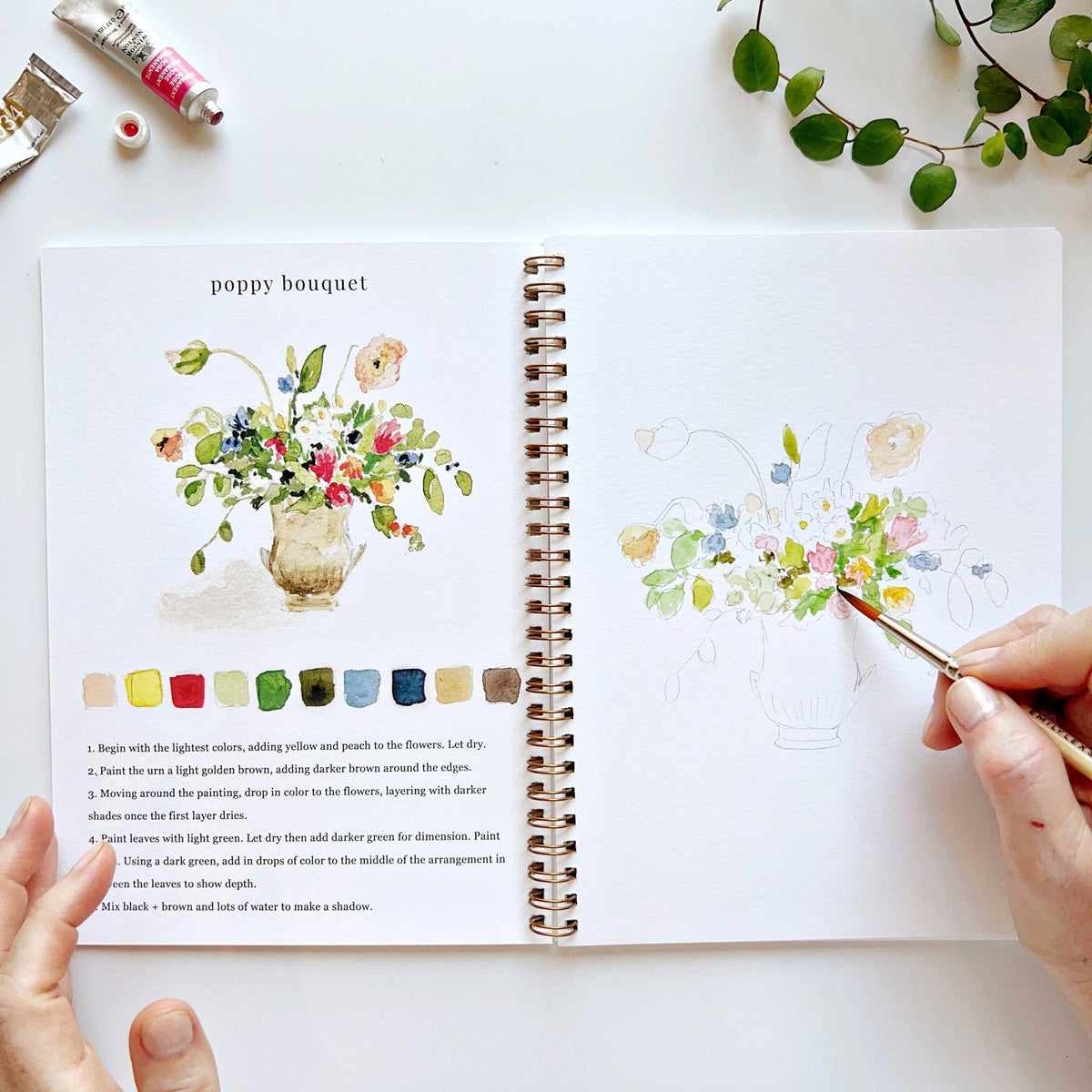 Floral watercolor workbook displayed alongside garden blooms, ceramic vessels, watercolor supplies, and natural textures, creating an inspiring creative workspace.