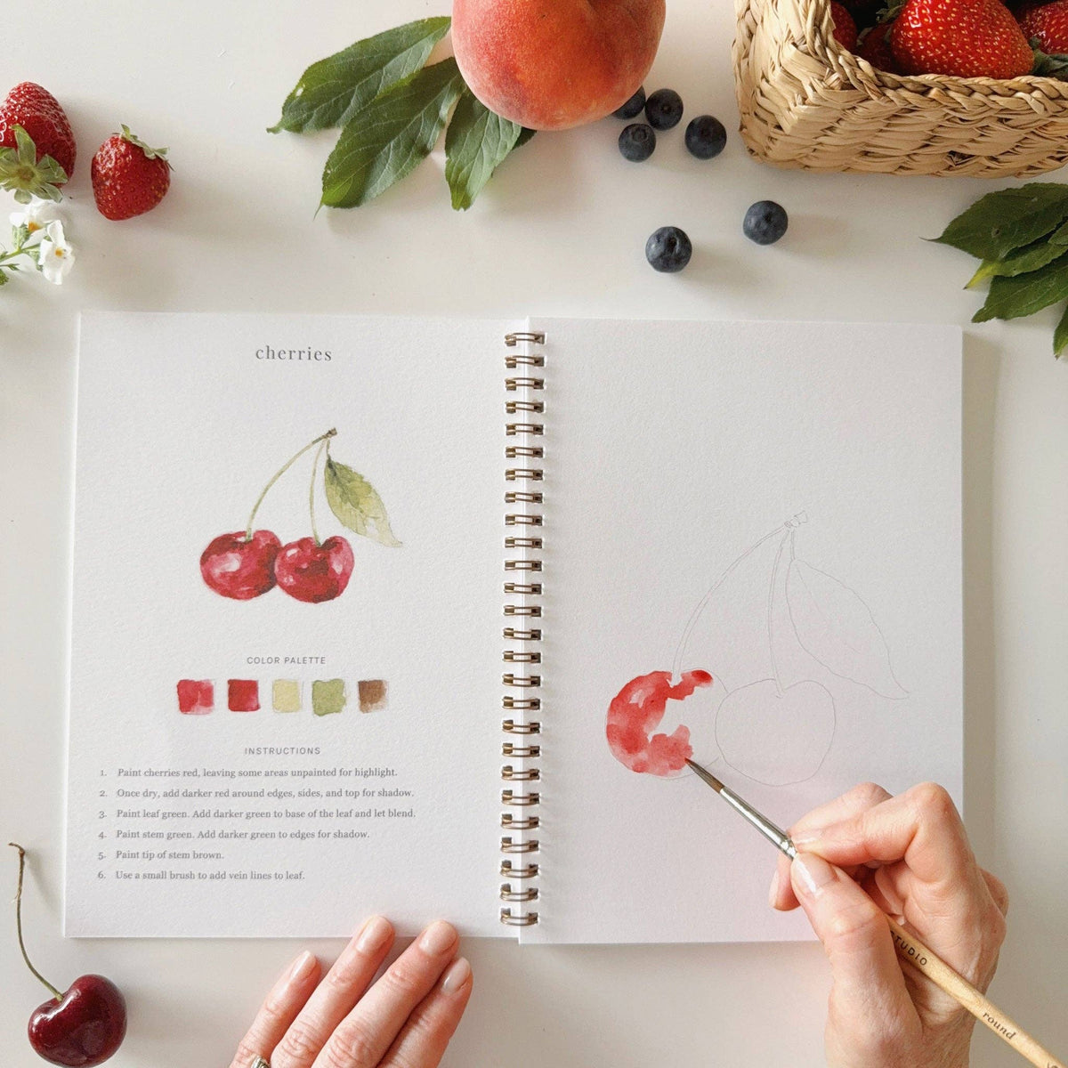 Open watercolor workbook displaying fruit illustrations with premium watercolor paper and easy-to-paint outlines.