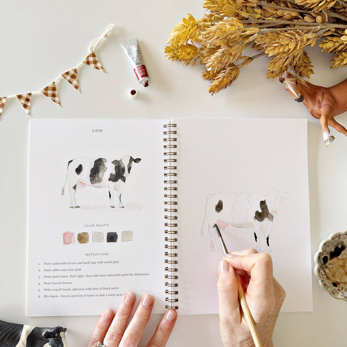 Open watercolor workbook displaying farm-themed illustrations on premium watercolor paper with easy-to-follow painting guidance.