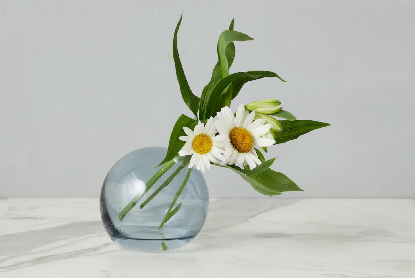 Blue glass vase with white flowers and green leaves on a light surface.