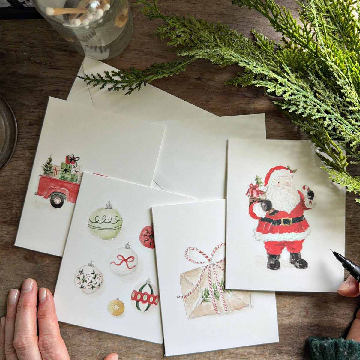 Four illustrated Christmas notecard designs by Emily Lex featuring Santa, a festive envelope, ornaments, and a red truck carrying gifts.