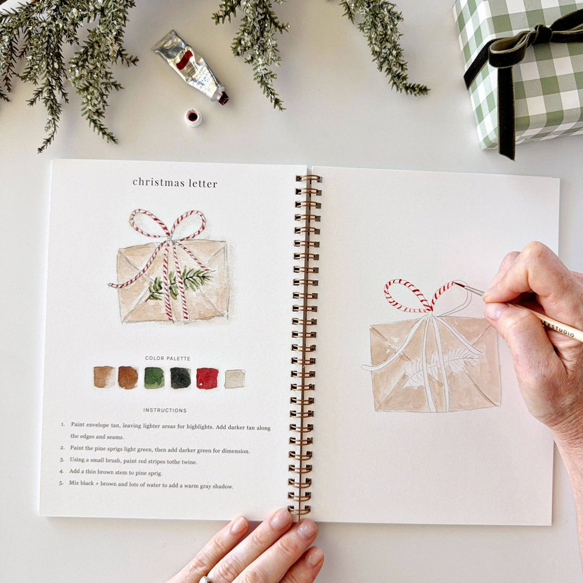 Open Christmas watercolor workbook showing a finished painting example, step-by-step instructions, and an outlined sketch.