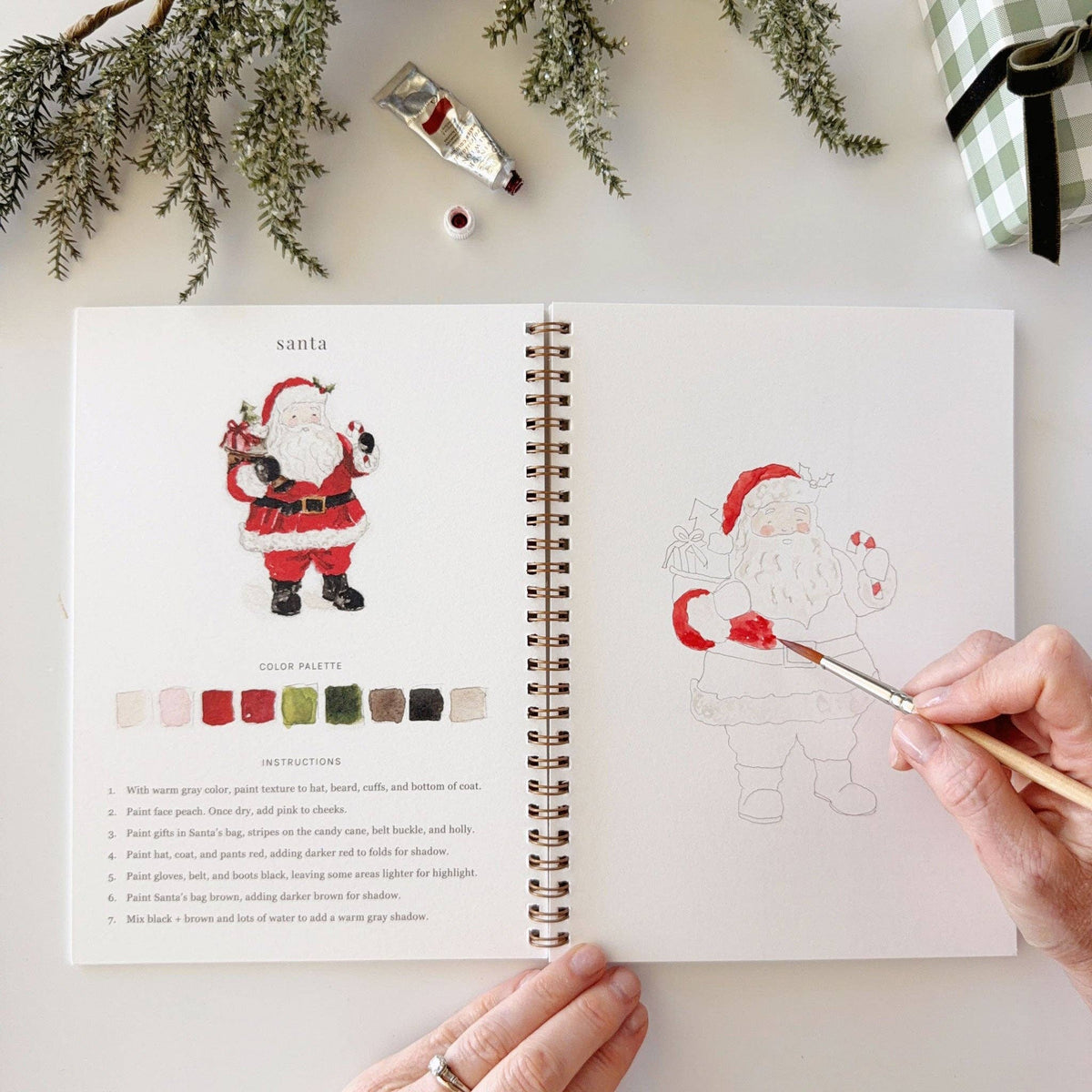 Emily Lex Christmas watercolor workbook open on a table with watercolor paints and brushes for a relaxing holiday painting activity.