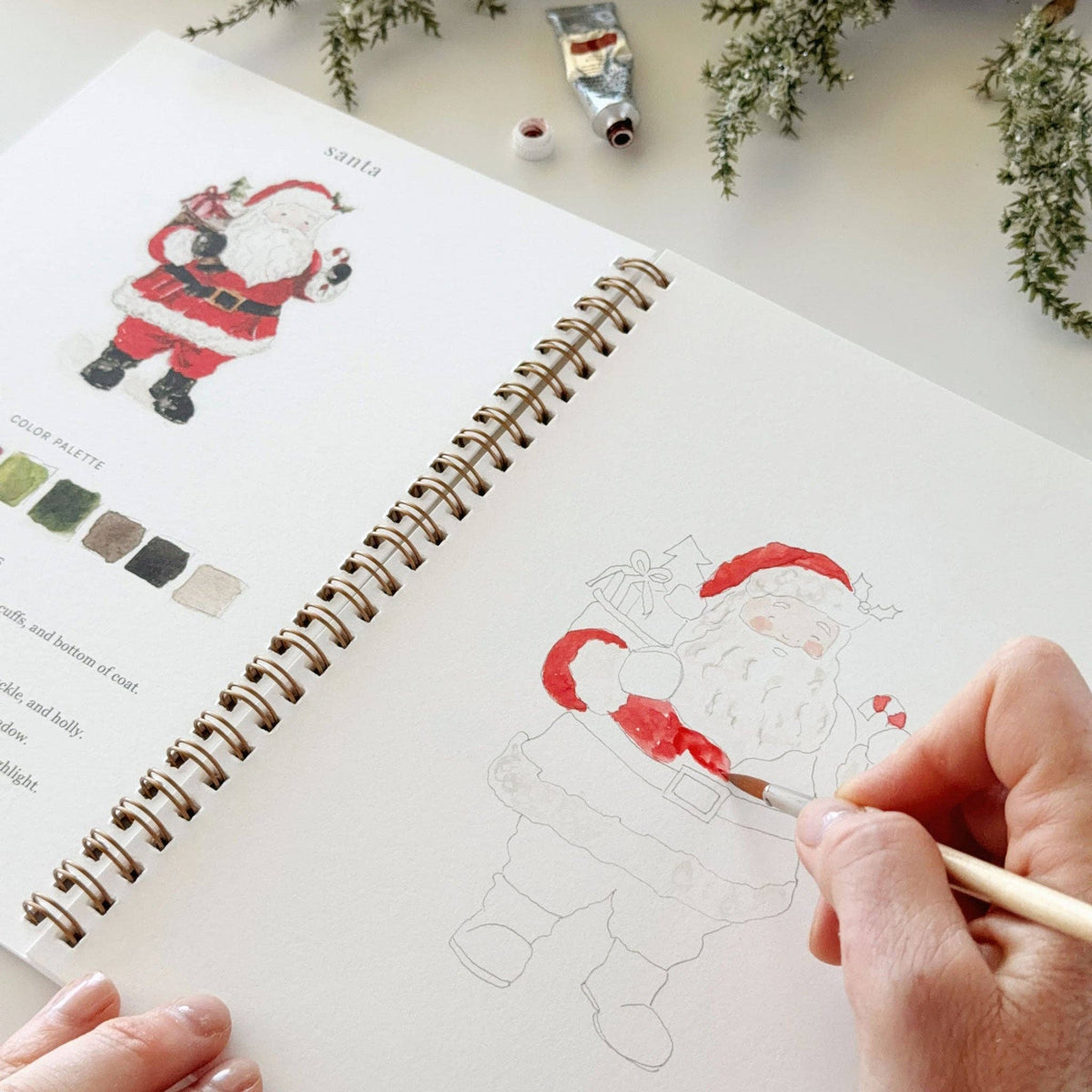 Emily Lex Christmas watercolor workbook open on a table with watercolor paints and brushes for a relaxing holiday painting activity.