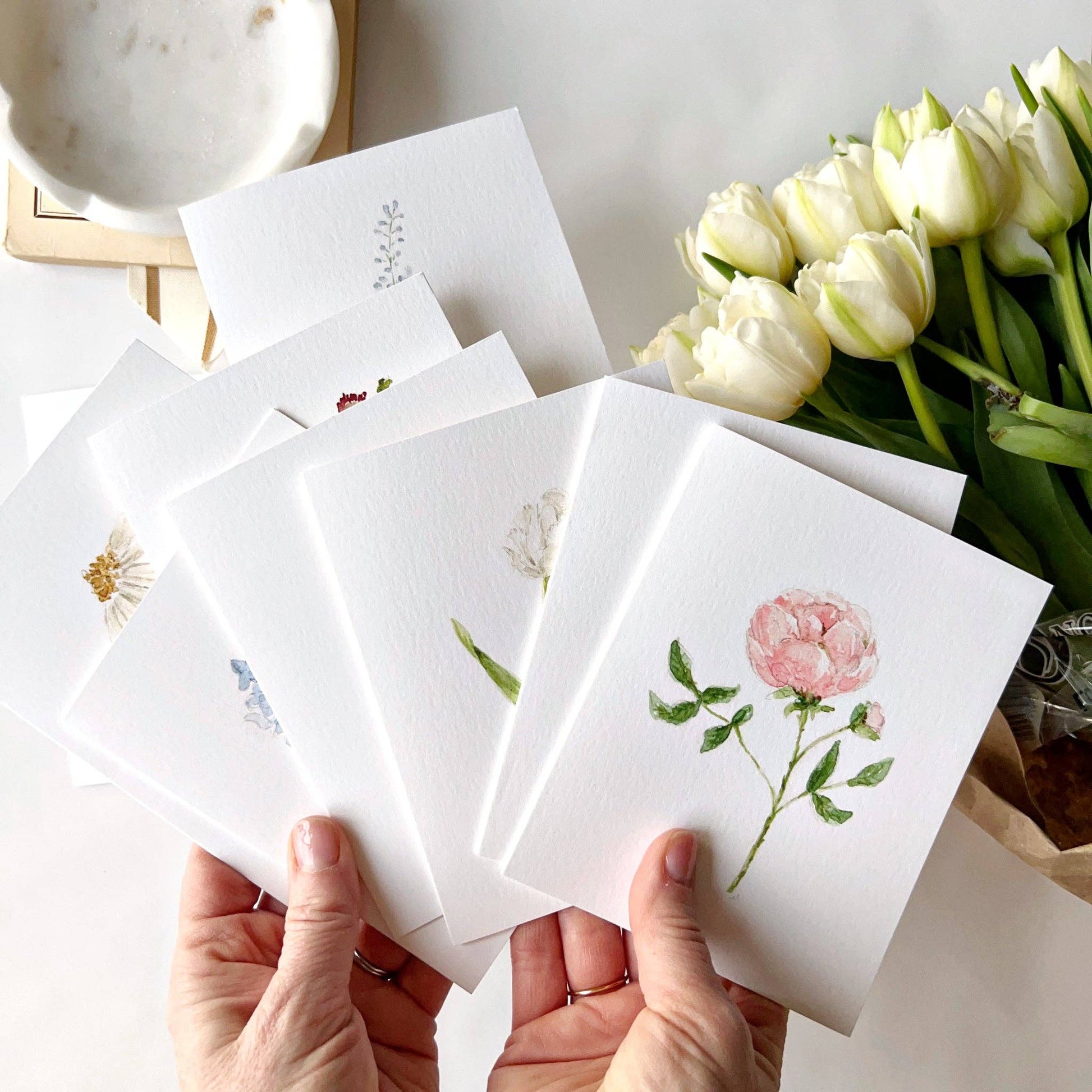 Assorted botanical notecard set displayed on a writing desk with fresh garden flowers, envelopes, and a fountain pen.