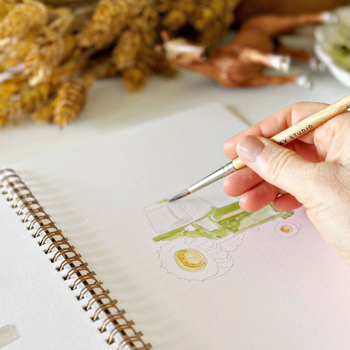 Farm-inspired watercolor workbook displayed with vintage gardening tools, linen textiles, watercolor supplies, and natural textures, creating a warm and inviting creative workspace.