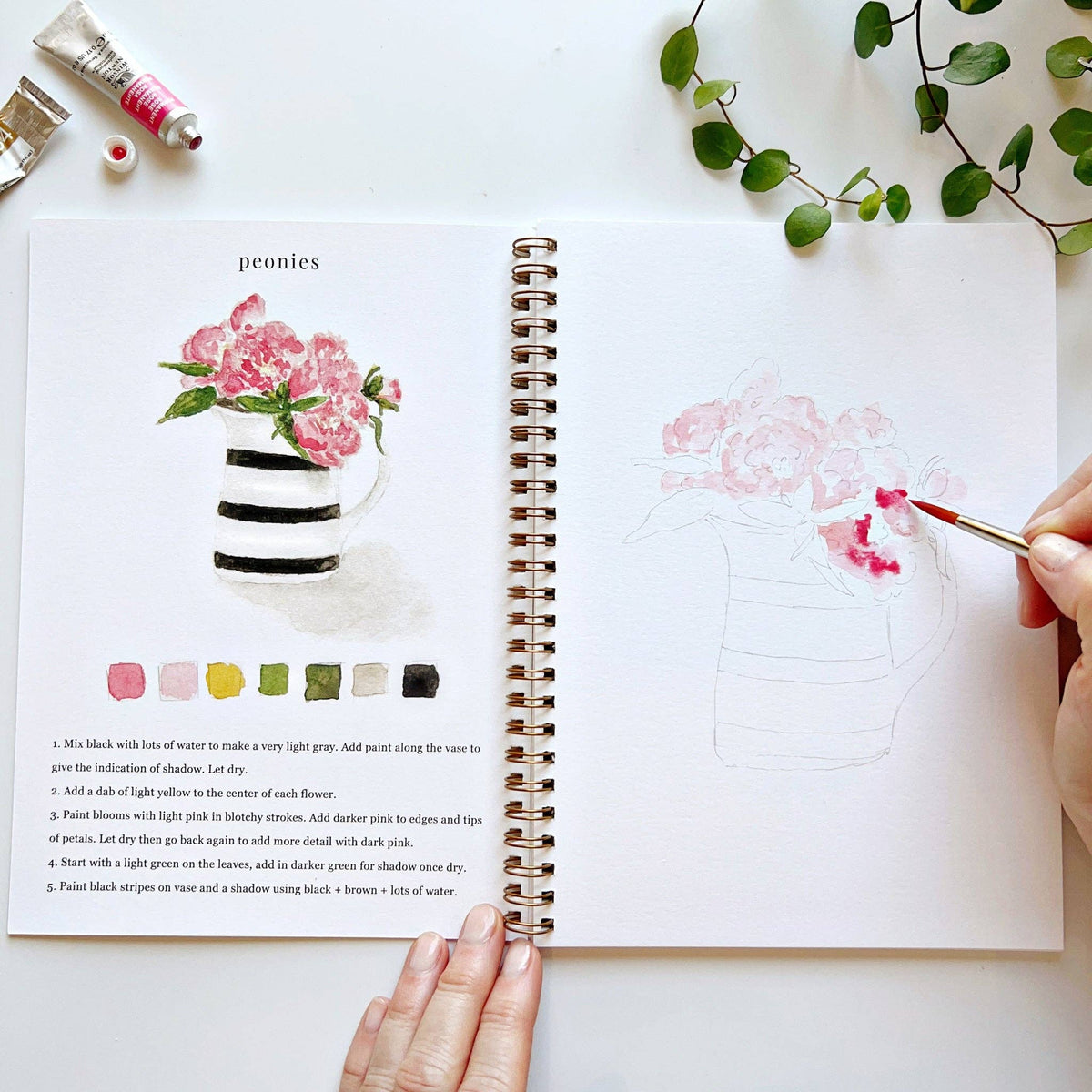 Open spiral-bound watercolor workbook displaying floral bouquet illustrations, step-by-step painting instructions, and premium watercolor paper.