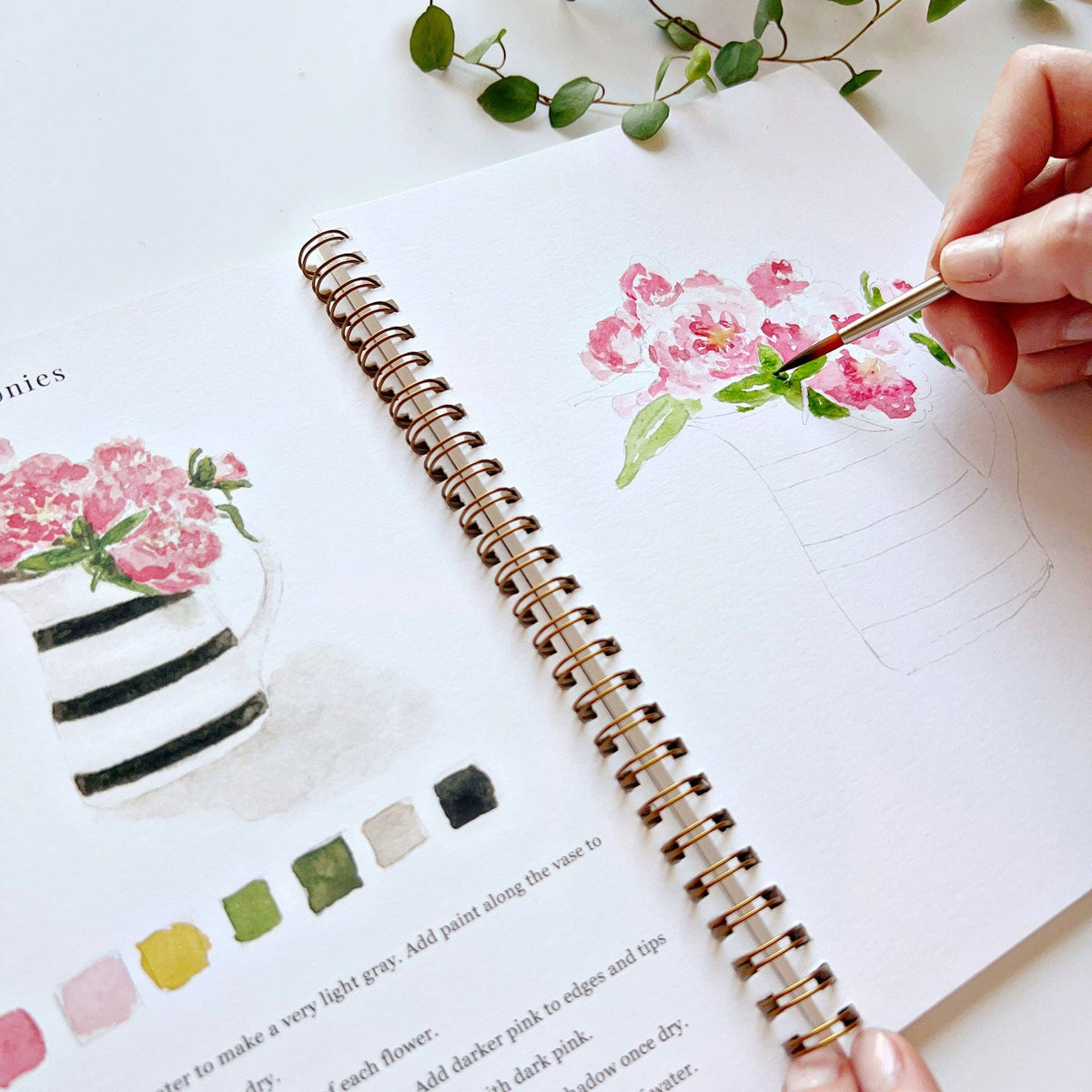 Open spiral-bound watercolor workbook displaying floral bouquet illustrations, step-by-step painting instructions, and premium watercolor paper.