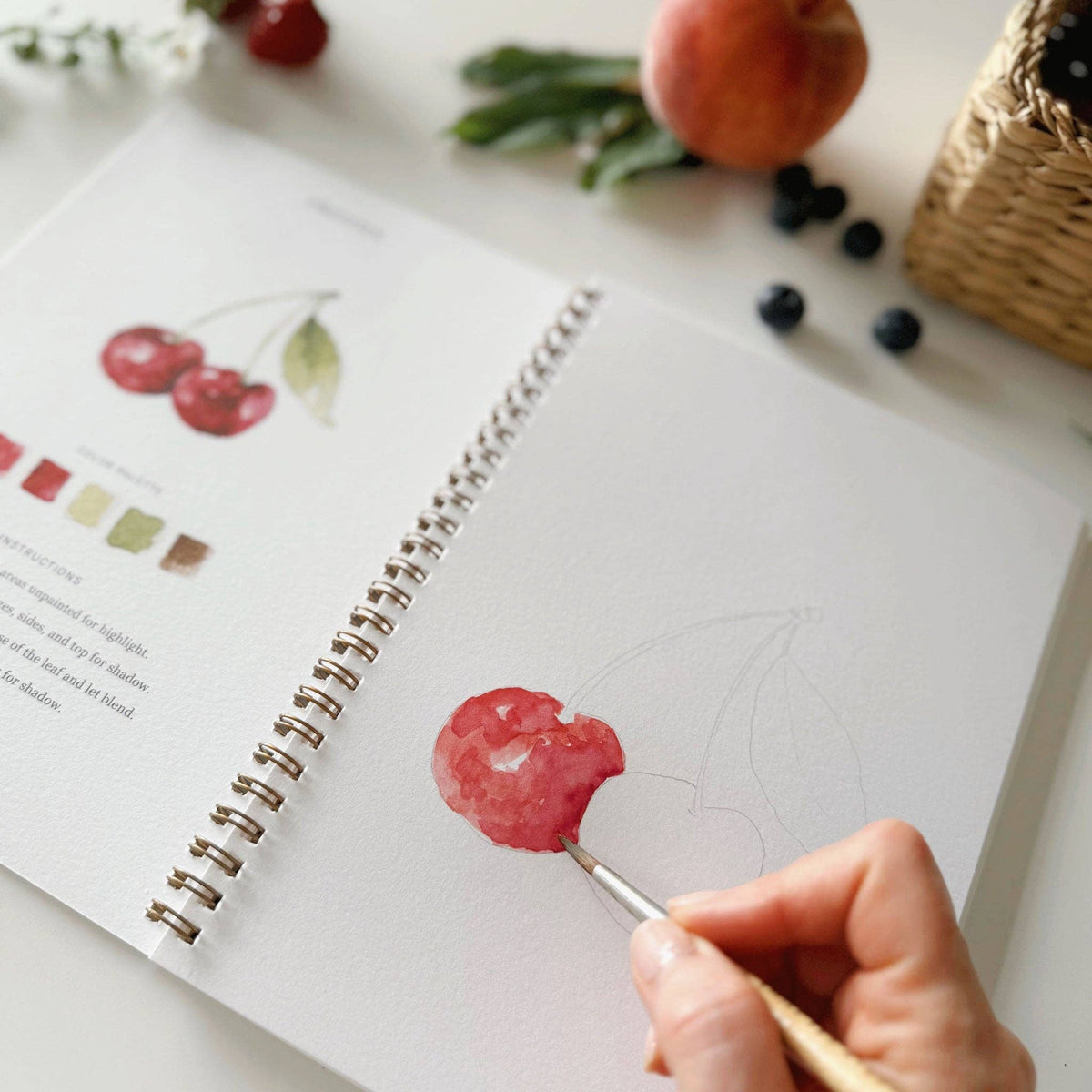Fruit watercolor workbook styled with watercolor paints, brushes, and fresh citrus on a bright creative workspace.