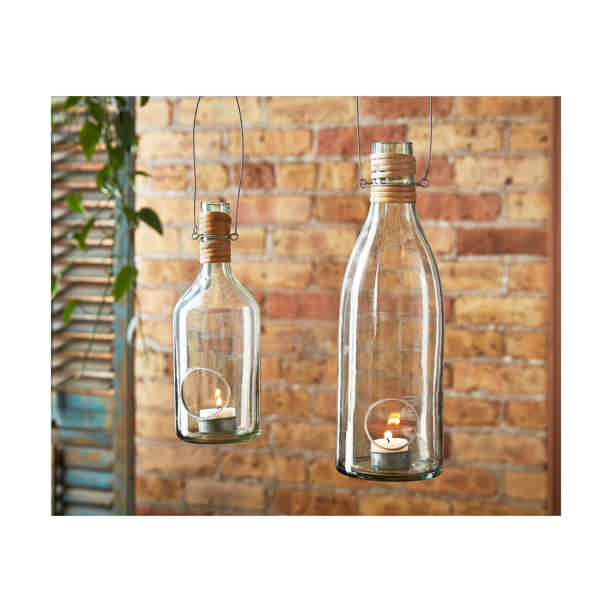 Two glass bottles with candles inside, hanging against a brick wall.