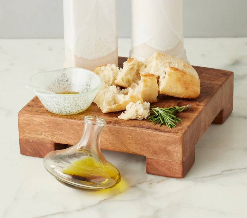 Wooden board with bread, cheese, and a glass container on a marble surface