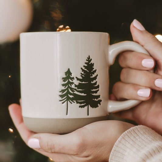 Cream stoneware coffee mug featuring a green evergreen tree design, speckled glaze, and exposed natural stoneware base.