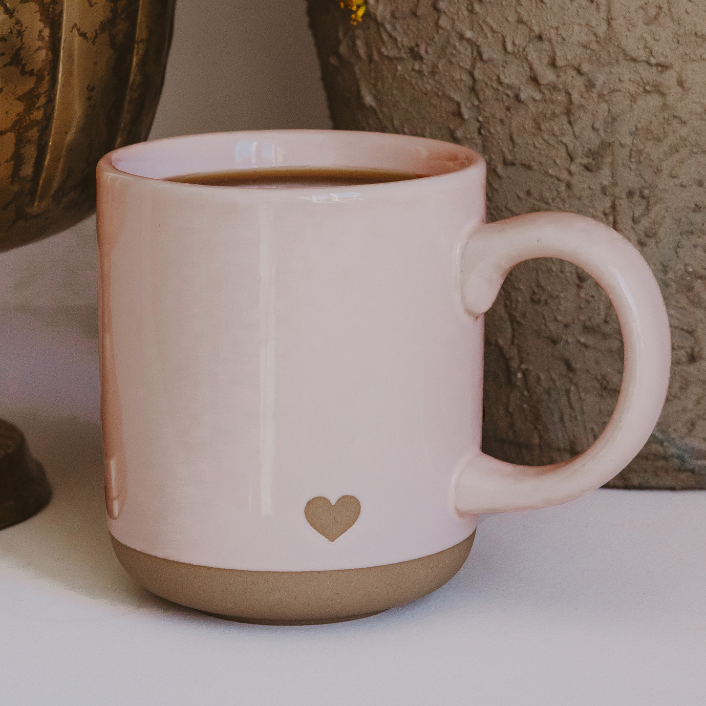 Light pink stoneware coffee mug with a heart design on both sides, a speckled glaze, and a natural exposed stoneware base.