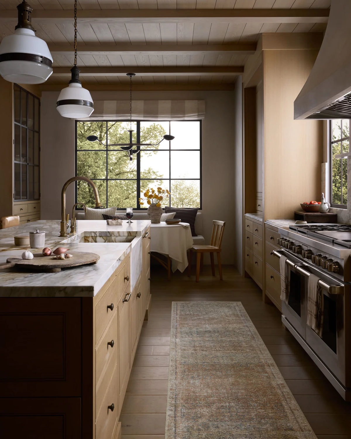 A wide, bright kitchen interior with light wood cabinetry, a marble countertop, a large window with outdoor greenery visible, a dining nook, a stainless steel gas range, pendant lights, and a long patterned runner rug on the floor.