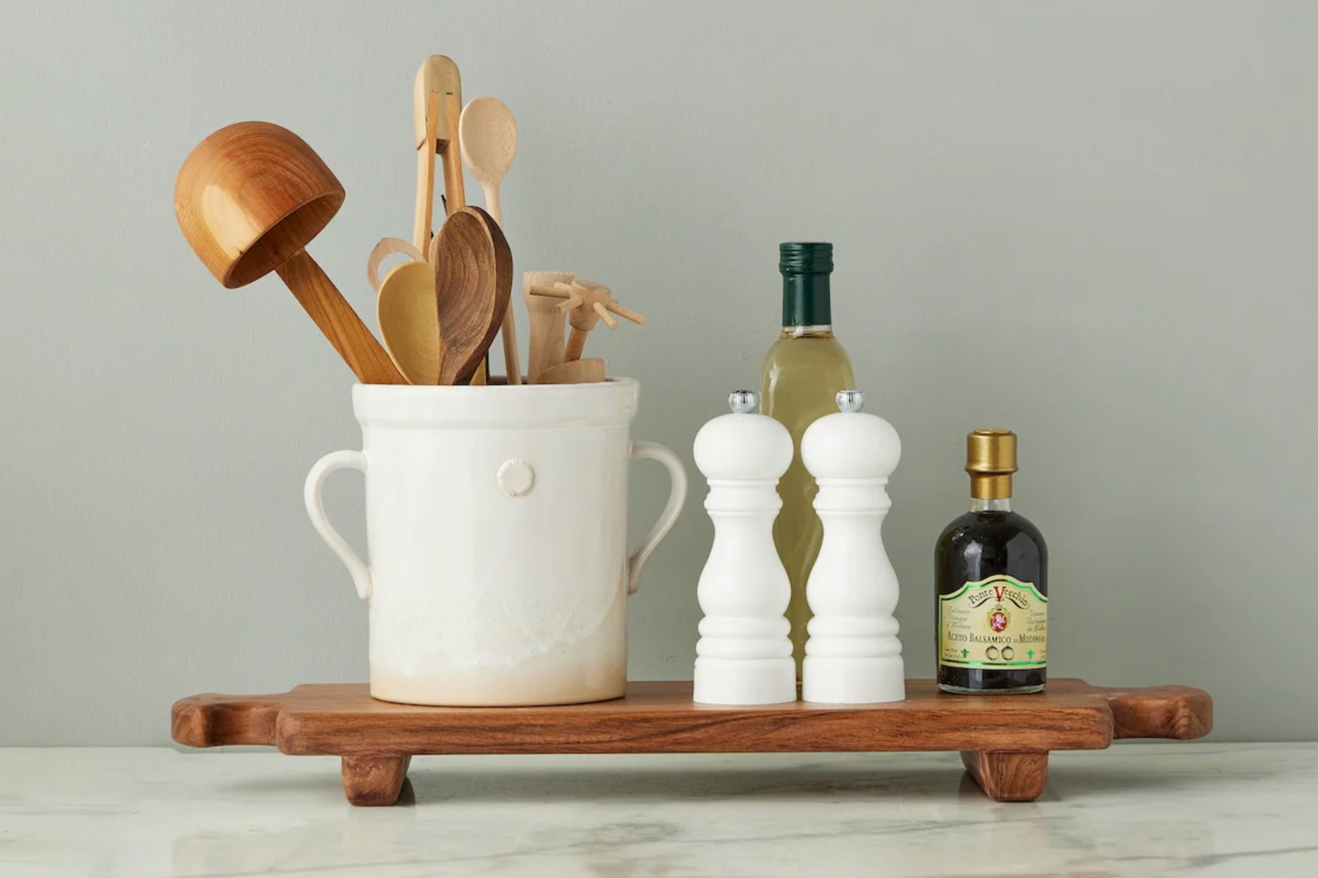 Wooden utensils in a white container on a wooden tray with bottles and salt and pepper shakers.