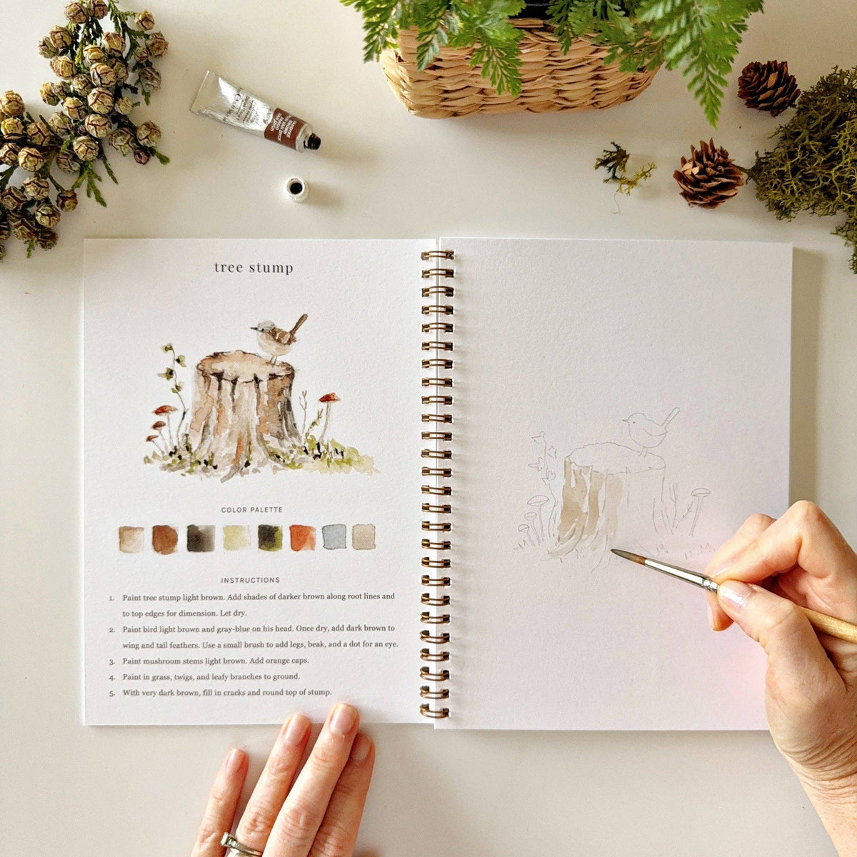 Woodland watercolor workbook displayed with natural textures, wildflowers, and art supplies, creating a peaceful and inspiring creative setting.
