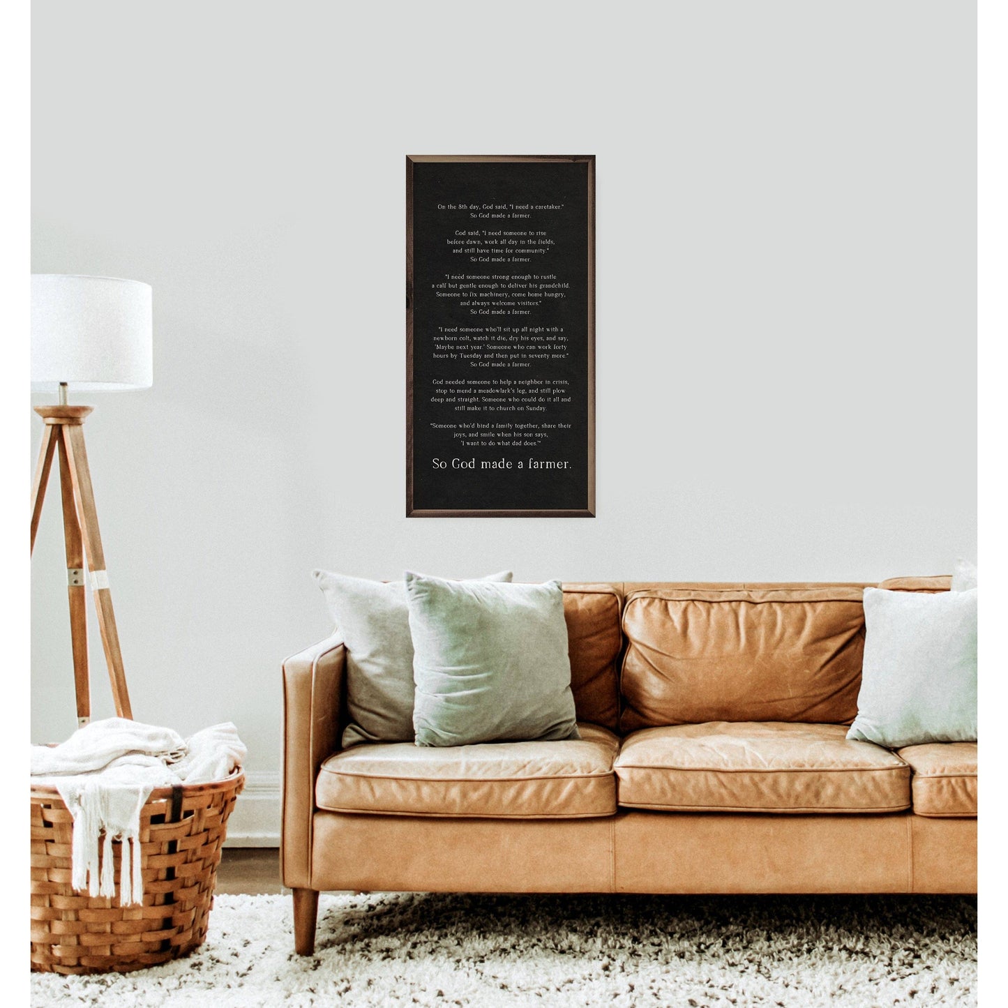 A framed black wall art print with white text hangs above a tan leather sofa in a styled living room with pillows, a floor lamp, and a basket on a rug.