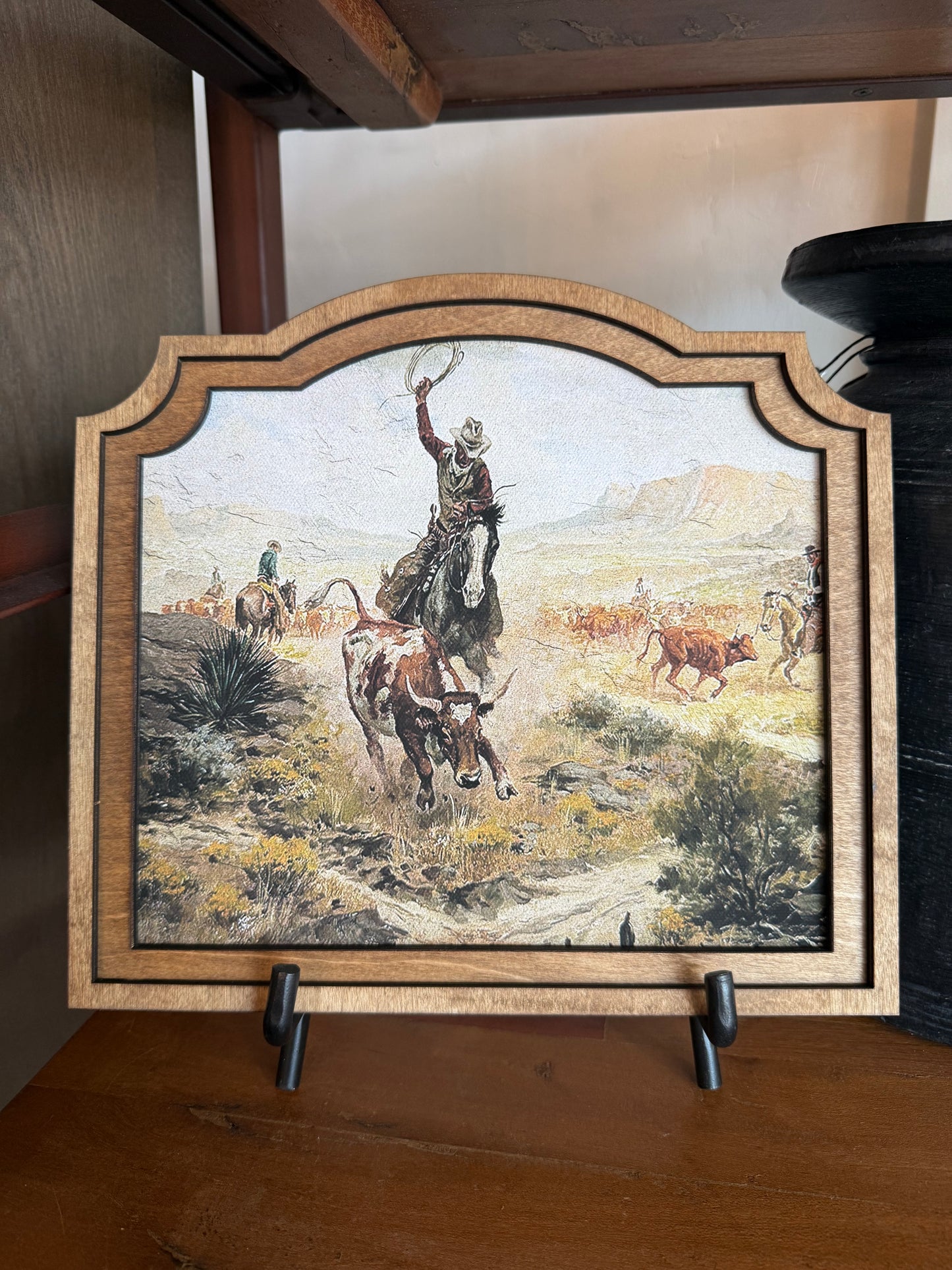 A framed western-themed art print on a wooden shelf, showing a cowboy riding a horse with other cattle and riders in the background. The piece has an arched top and a light wood frame, displayed on small black stands.