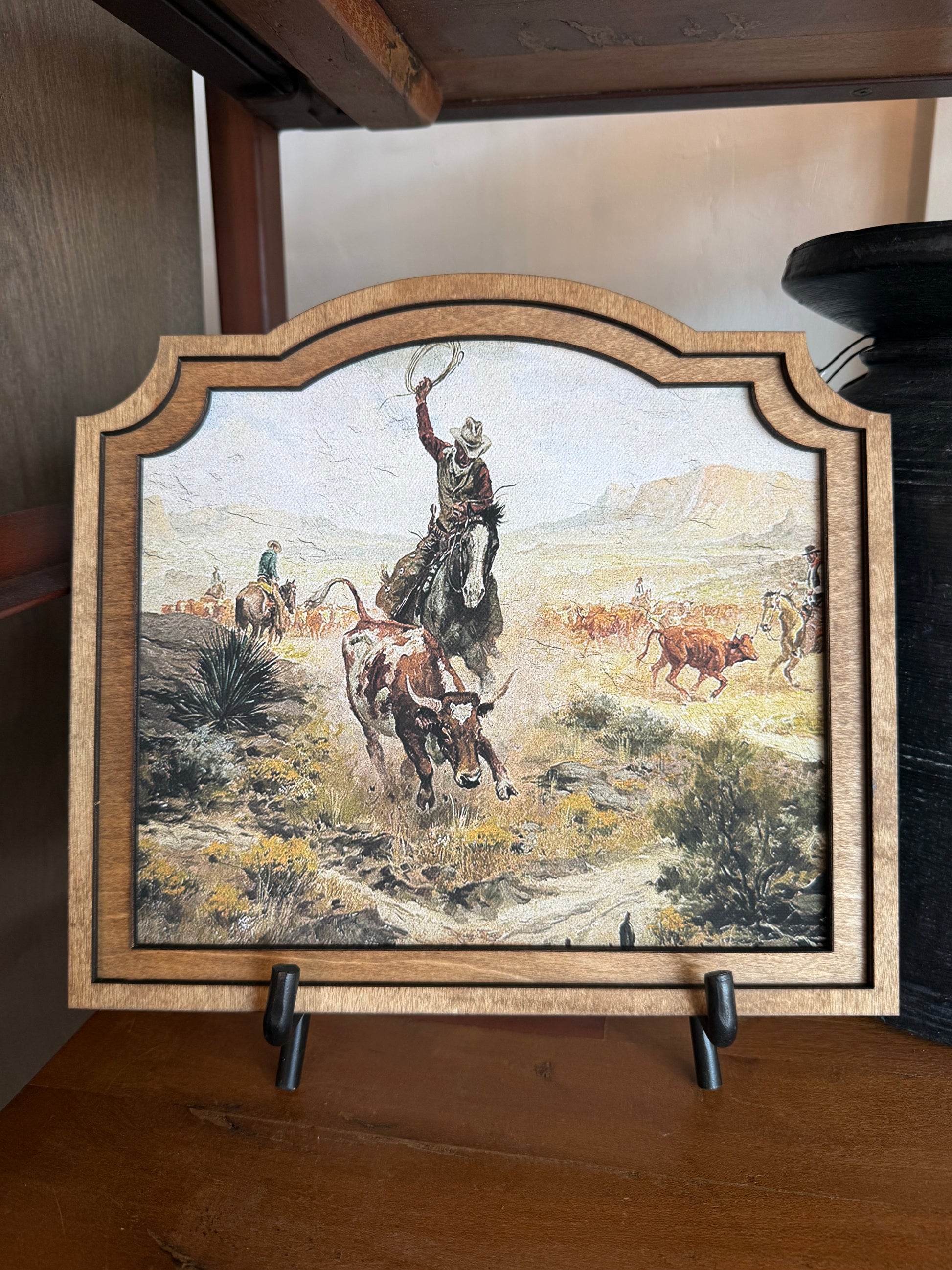 A framed western-themed art print on a wooden shelf, showing a cowboy riding a horse with other cattle and riders in the background. The piece has an arched top and a light wood frame, displayed on small black stands.