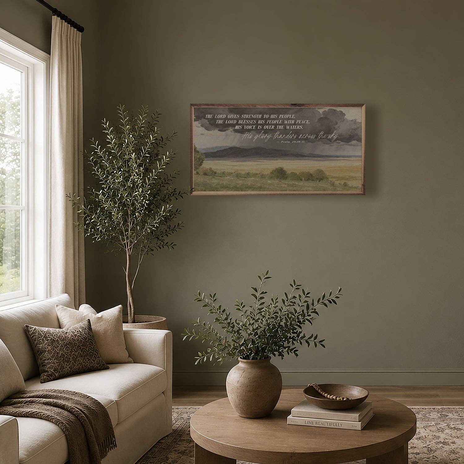 A framed landscape art print with a Bible verse hangs on a sage green wall in a styled living room with a beige sofa, a wooden coffee table, and potted plants.