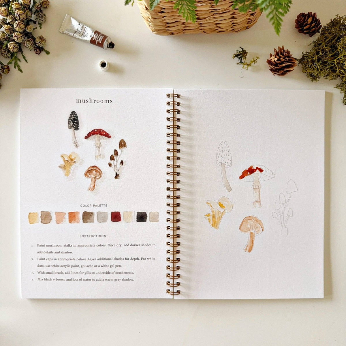 Woodland watercolor workbook styled with watercolor paints, brushes, and fresh greenery on a bright creative workspace.