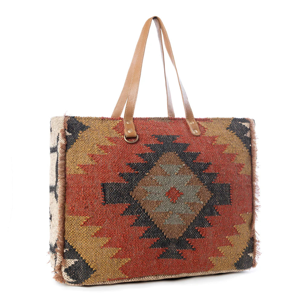 Kaliko Hill Weekender Bag featuring a woven tribal-inspired rug textile, caramel leather straps, and a spacious zip-top design.